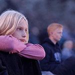 Ung blond pige sidder med armene omkring knæene i skumringen under teaterforestilling ved Minach Teatret.